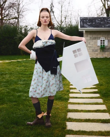 Adult female model in light blue dress holds white toy house outdoors on grass with small outbuilding. Fashion editorial image.