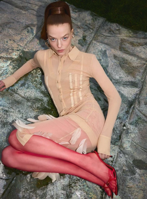 A fair-skinned woman with red hair tied high poses in a sheer beige collared dress, reddish tights, and heels on a textured surface.