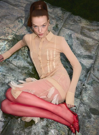 A fair-skinned woman with red hair tied high poses in a sheer beige collared dress, reddish tights, and heels on a textured surface.