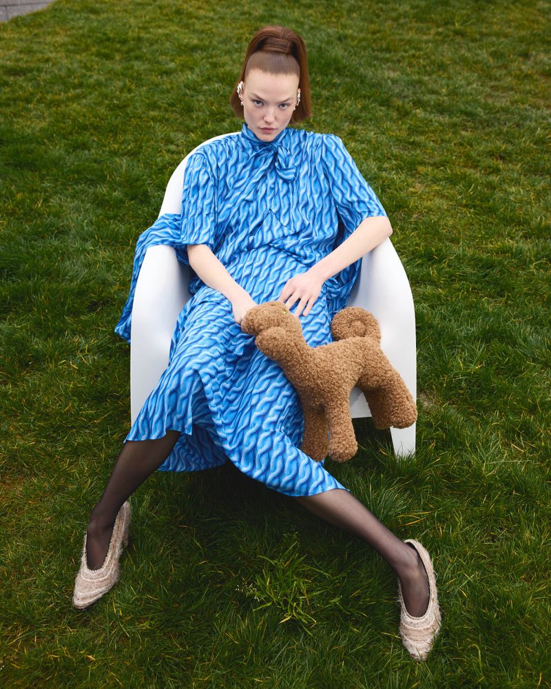 A young adult female model with auburn hair poses in a chair outdoors on a grassy surface, wearing a blue patterned dress and holding a teddy bear.