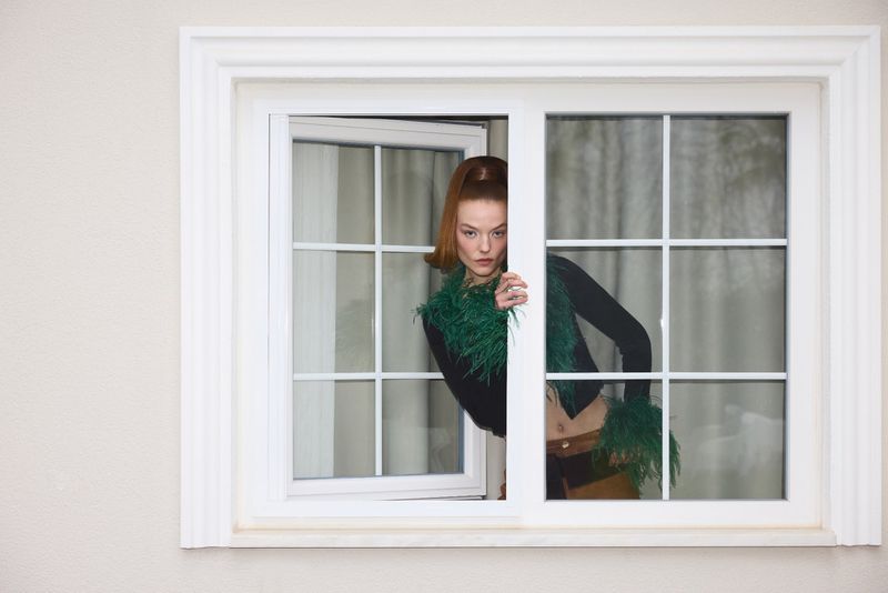 A young, fashionable adult woman with red hair is framed within a white window, creating a candid and stylish moment indoors.