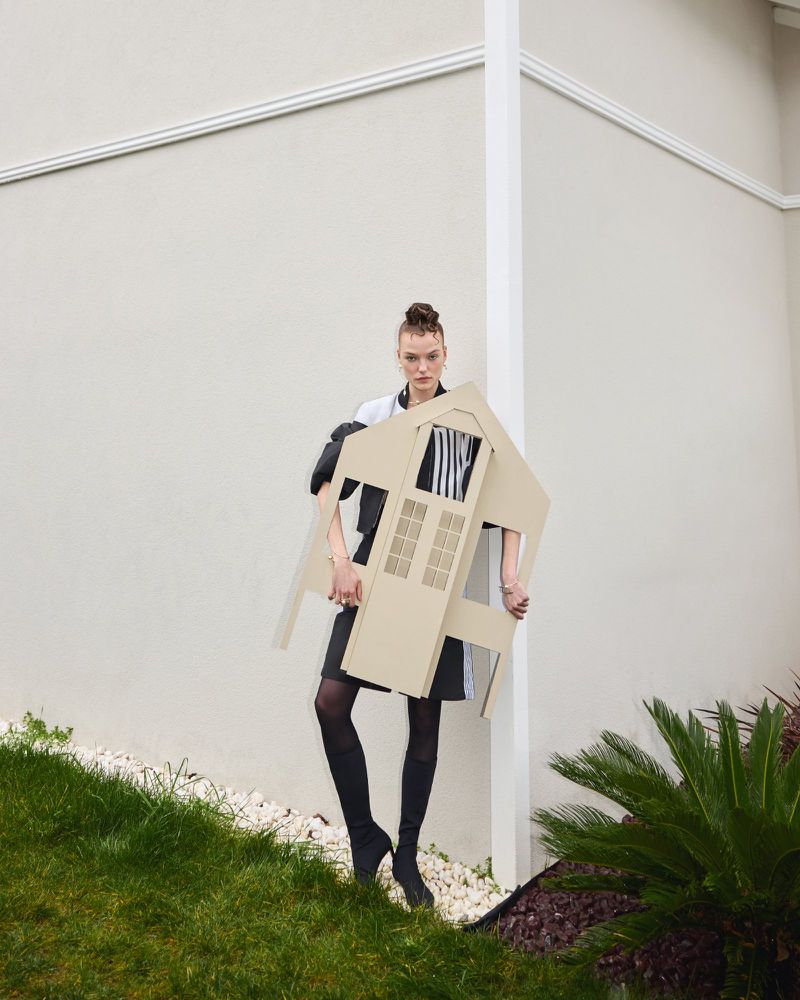 Adult female poses outdoors, holding house-shaped prop. She wears boots, a black skirt, and a jacket. Green grass and a building are visible.