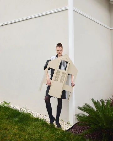 Adult female poses outdoors, holding house-shaped prop. She wears boots, a black skirt, and a jacket. Green grass and a building are visible.