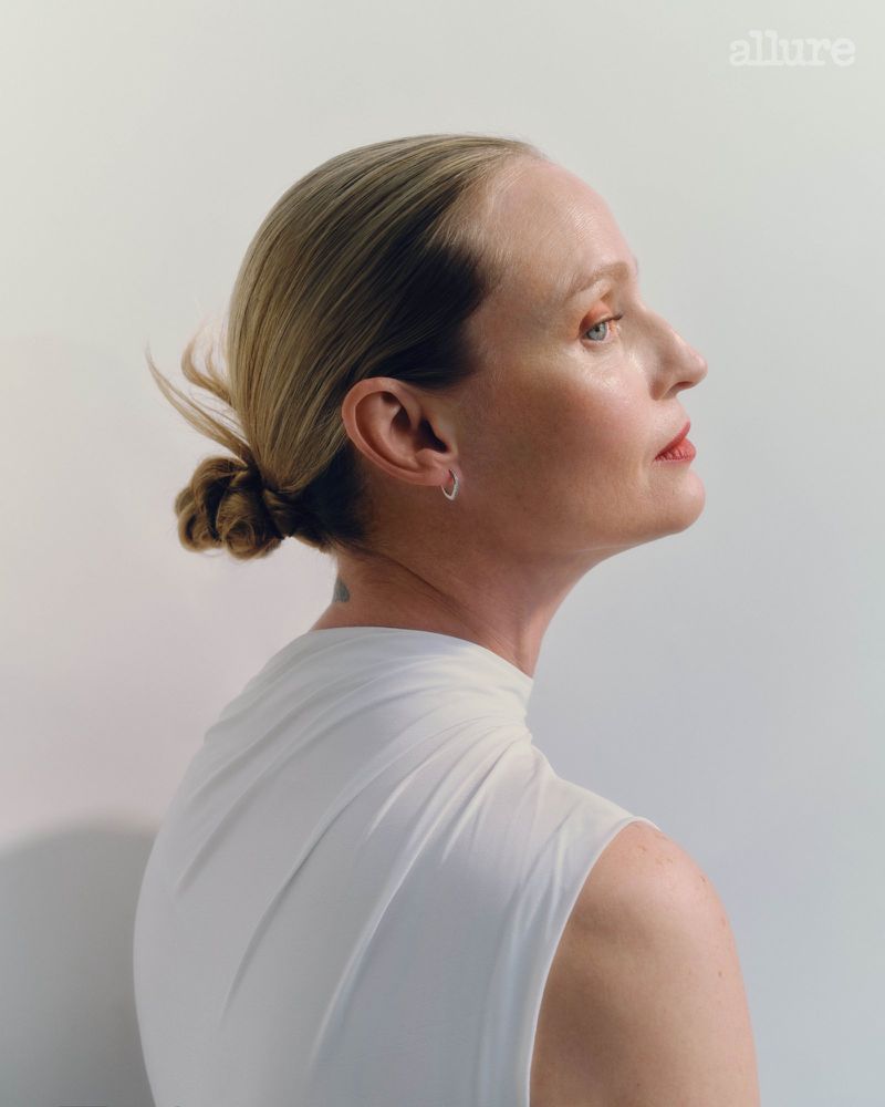 Portrait of a blonde adult female in a draped white top against a plain backdrop, styled for Allure magazine with a minimalist aesthetic.