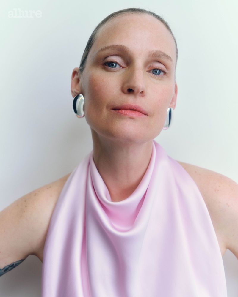 Elegant portrait of an adult woman with blue eyes and silver earrings. She wears a sleeveless light-pink satin top against a white backdrop.