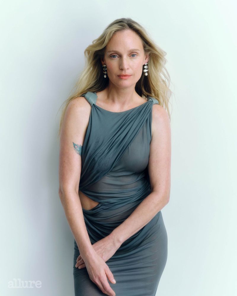 Blond woman in a grey dress poses against a white wall wearing pearl earrings in a beauty shot with soft, neutral lighting.