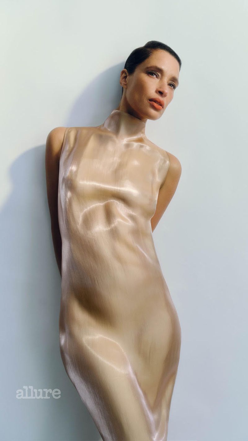 A fair-skinned woman with short dark hair poses in a shiny, tight-fitting golden dress against a white wall. Fashion editorial shot.