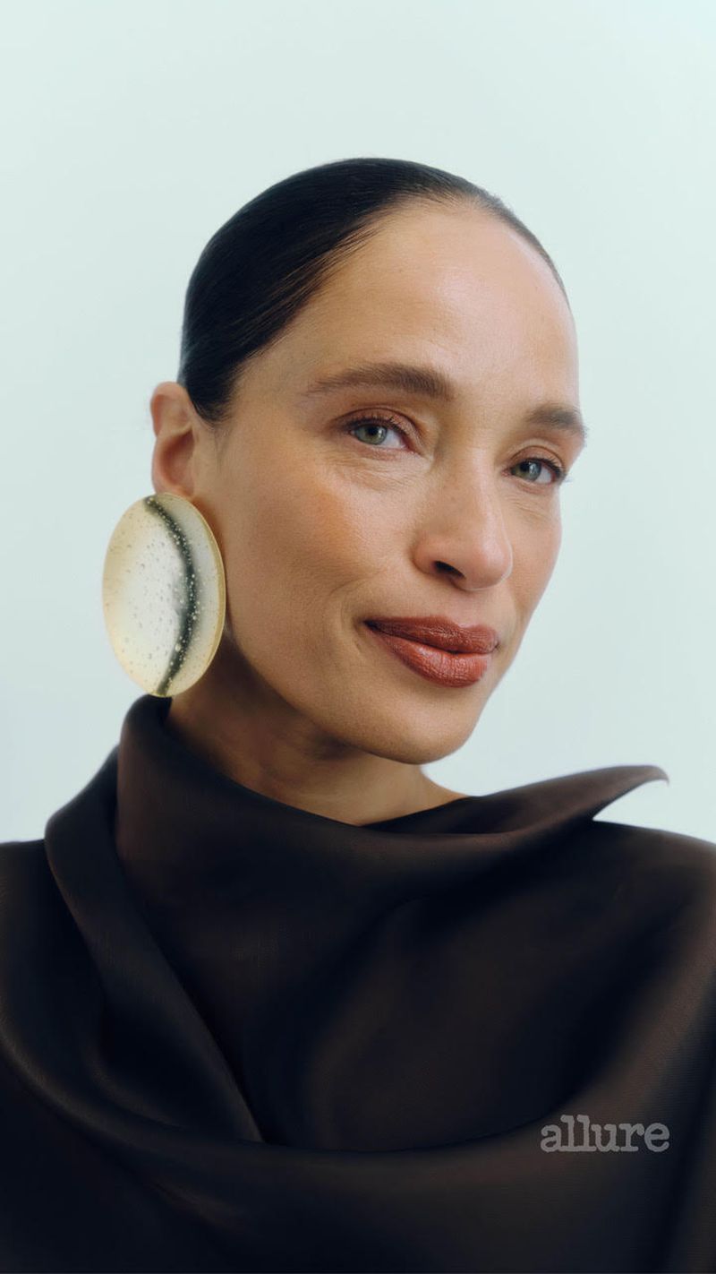 Close-up of an adult woman with dark hair and brown top wearing large round earrings against a light background. Beauty shot.