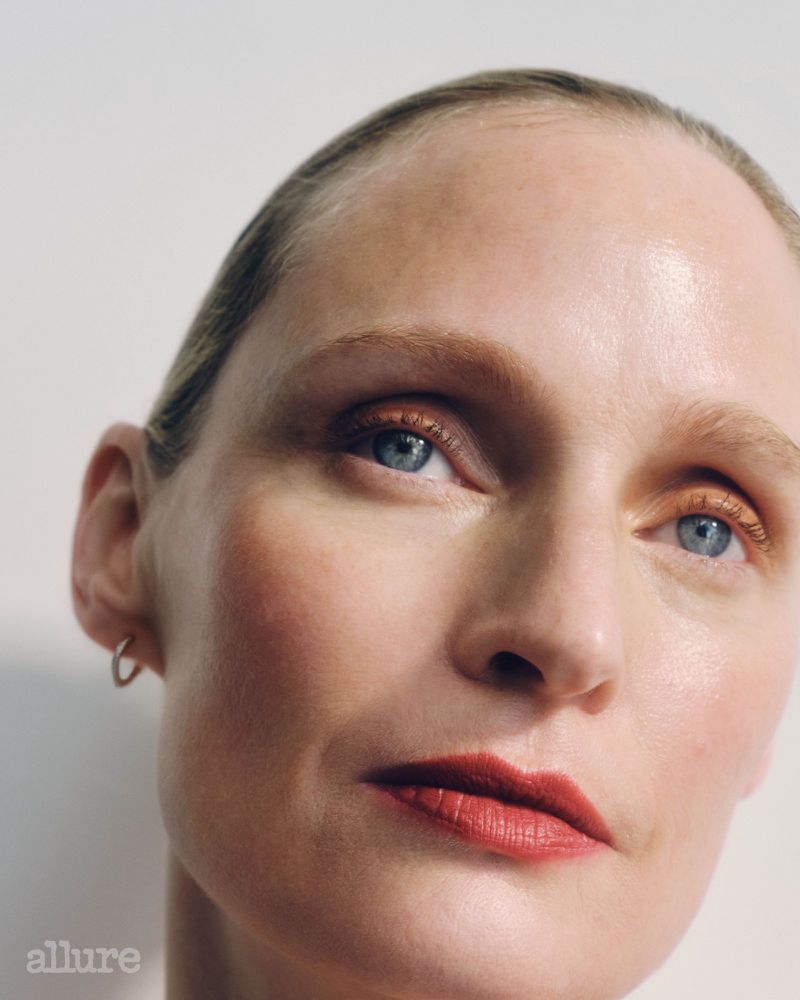 Close-up studio portrait of an adult woman with blonde hair, makeup, and gold hoop earring against a neutral background.