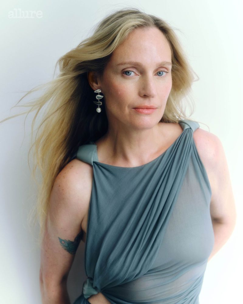 Portrait of a blonde woman with long hair wearing a draped gray-blue dress and statement earrings, posing against a white background.