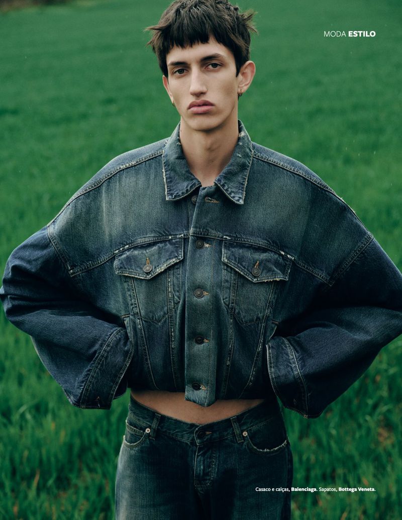 Close-up studio shot of a young adult man wearing a dark blue denim jacket and jeans posing in an outdoor grassy setting.