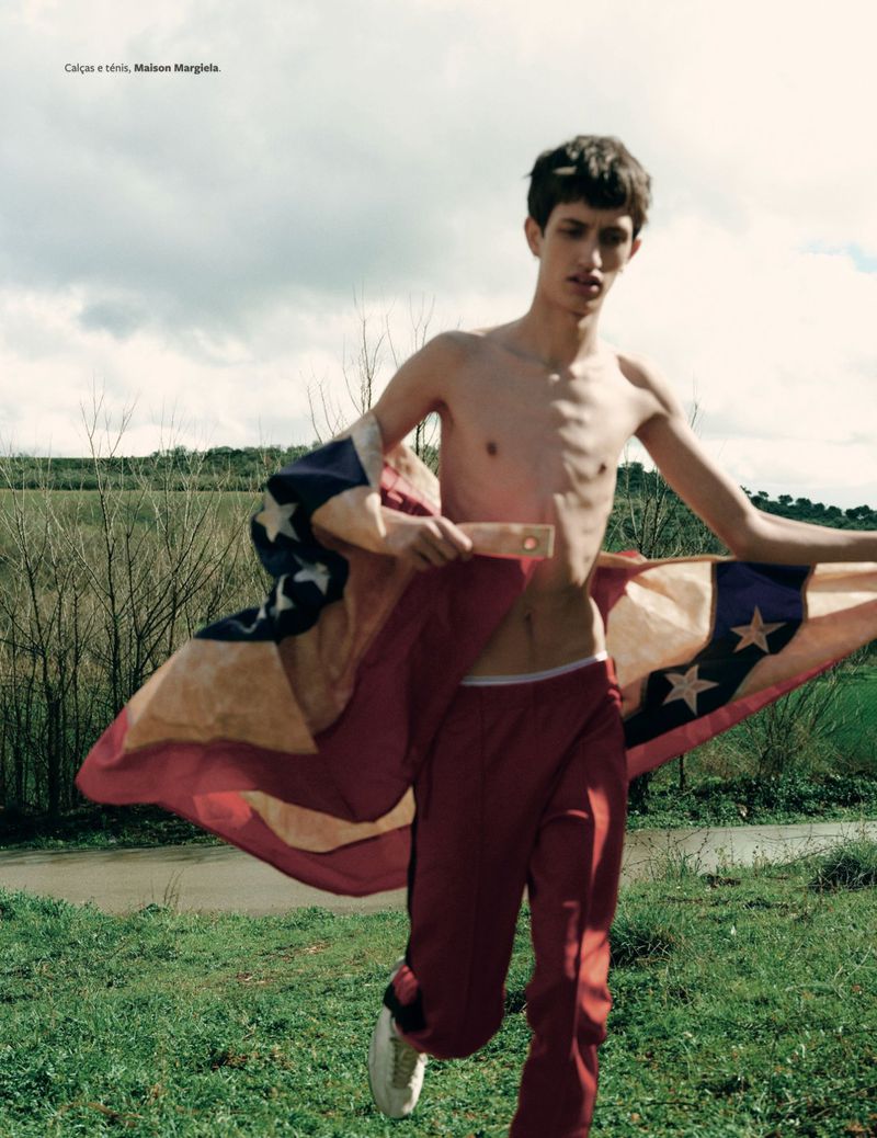 A young adult male runs in a field, draped in a tattered American flag, wearing red pants and white sneakers, under an overcast sky.