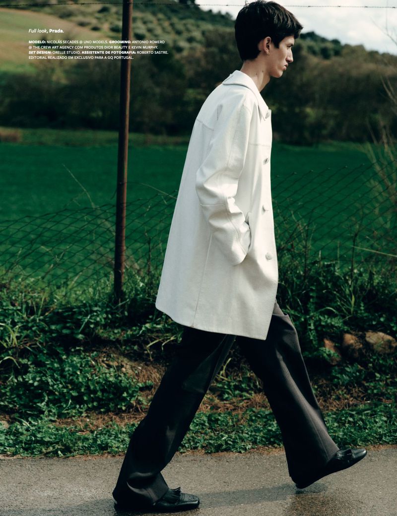 A light-skinned male model in a white overcoat and black pants walks on a paved path surrounded by a green landscape.