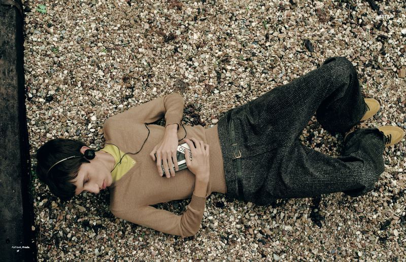 Young man lying on a gravel surface with headphones on, holding a walkman; wearing a tan sweater and dark pants, with yellow shoes.