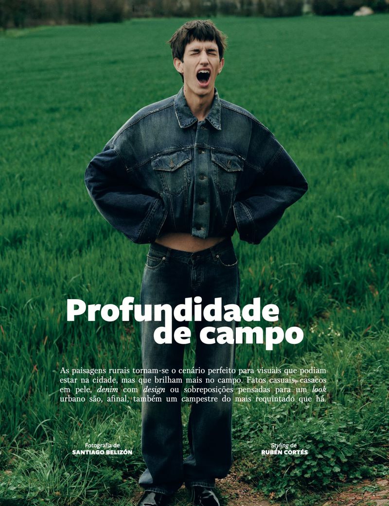 A young adult male model in a denim jacket and jeans stands in a field with green grass, looking up and shouting with his mouth open.