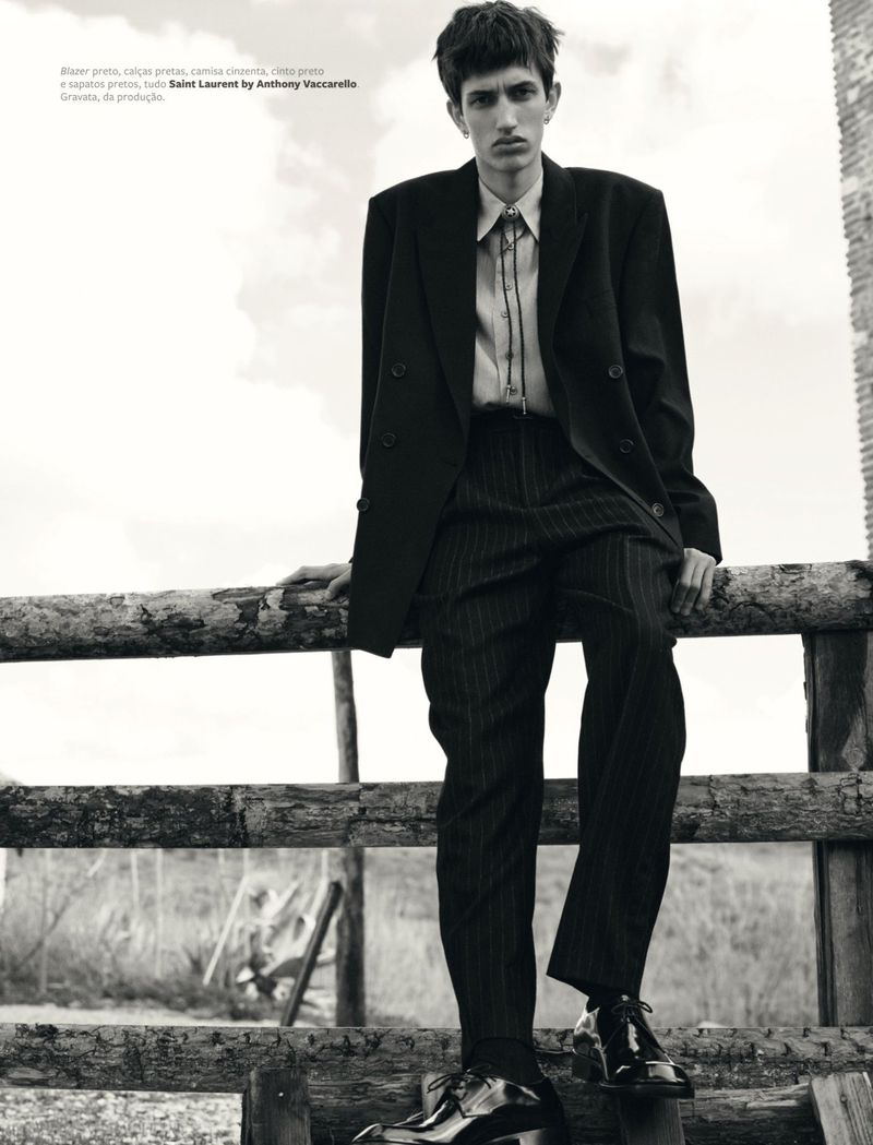 A male fashion model in a black suit and shiny black shoes poses against a wooden fence in a black and white fashion shot.