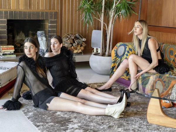 Three women in stylish black outfits pose in a retro interior with wood paneling, a fireplace, and a patterned couch, exuding fashion and style.