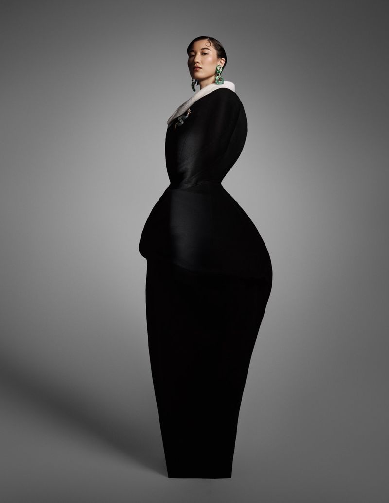 Elegant woman in a dark gown with a white collar and green earrings poses in a full shot against a solid gray background.
