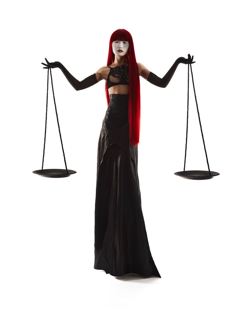 Full-length shot of a person in a black outfit and striking makeup, balancing a Libra scale against a bright white background.