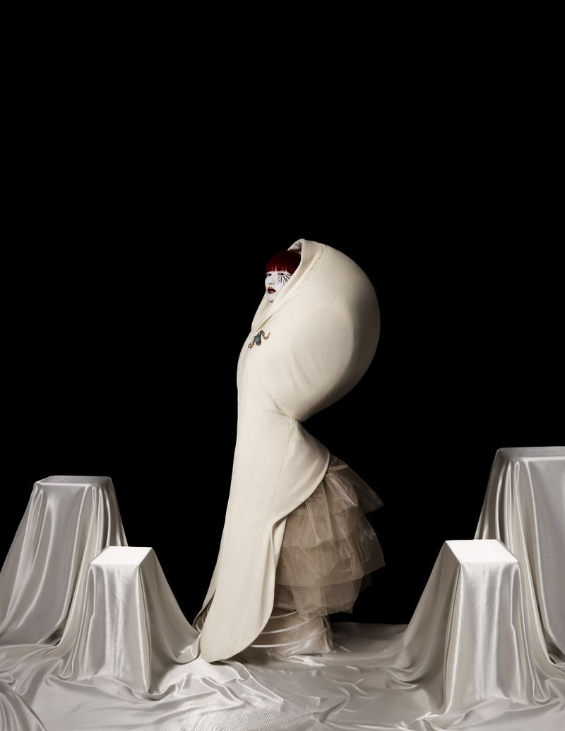 Person in sculptural white avant-garde dress with tulle, posing in a dark studio setting with draped pedestals.