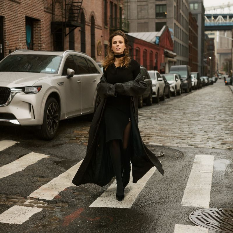 Stylish woman in a black dress and coat walks across a crosswalk in an urban setting, with cars and buildings in the background.