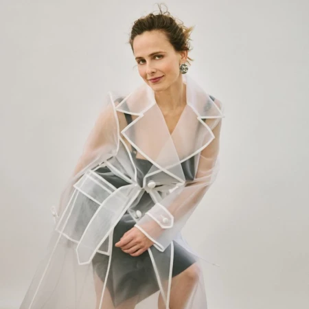 Portrait of a woman in a semi-sheer, white, trench coat with a cinched waist against a plain white backdrop in a studio environment.