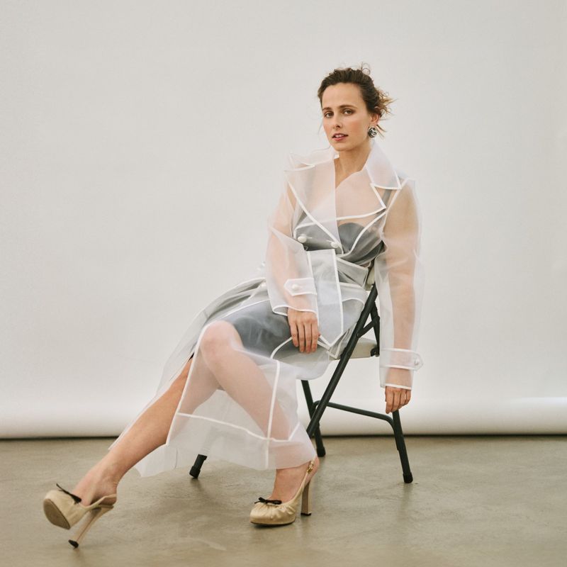 A fair-skinned adult woman seated, wearing a transparent coat and heels against a neutral backdrop, striking a fashion pose.