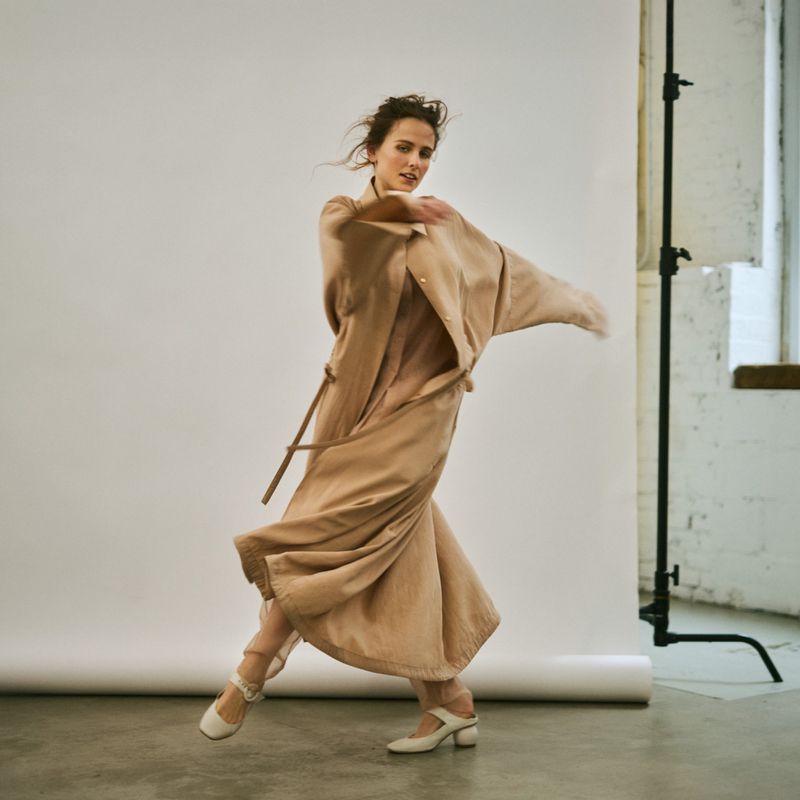 A woman in a beige outfit is captured mid-motion in a studio setting with a white backdrop and natural light from a nearby window.