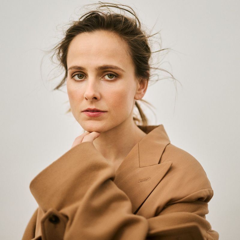 Portrait of a young adult female with fair skin and green eyes wearing a tan jacket, shot at eye-level against a light background.