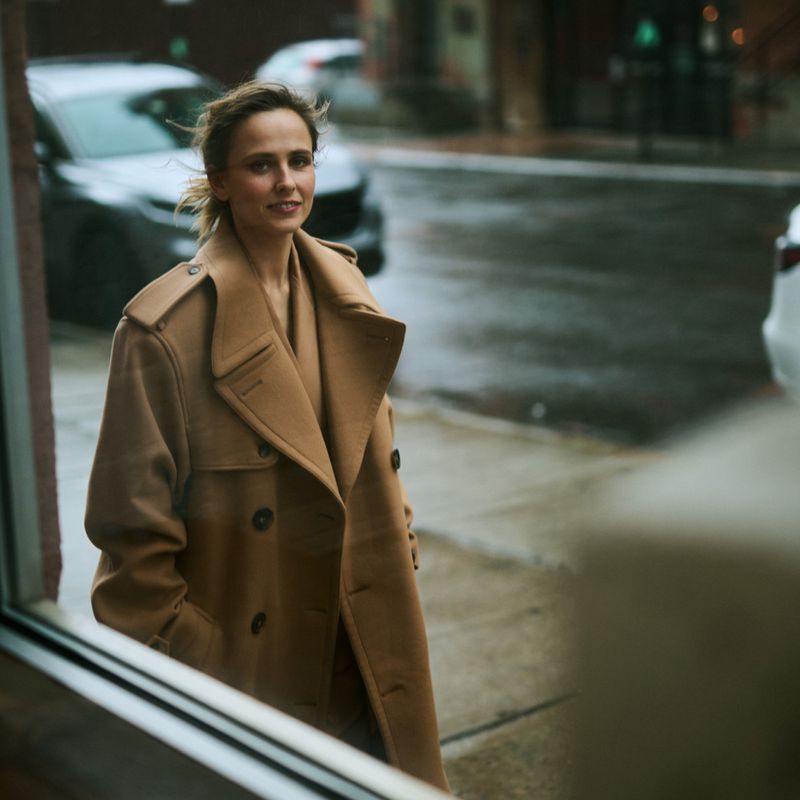 Woman in stylish tan coat standing on a city street corner near a building. She has shoulder-length hair and is smiling slightly.