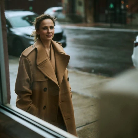 Woman in stylish tan coat standing on a city street corner near a building. She has shoulder-length hair and is smiling slightly.