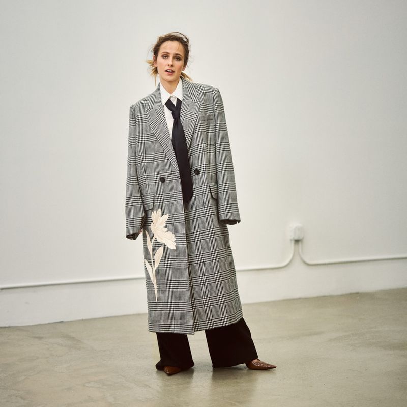 A stylish woman in a plaid coat, white collared shirt, and black tie confidently poses in a minimalist indoor setting with a concrete floor.