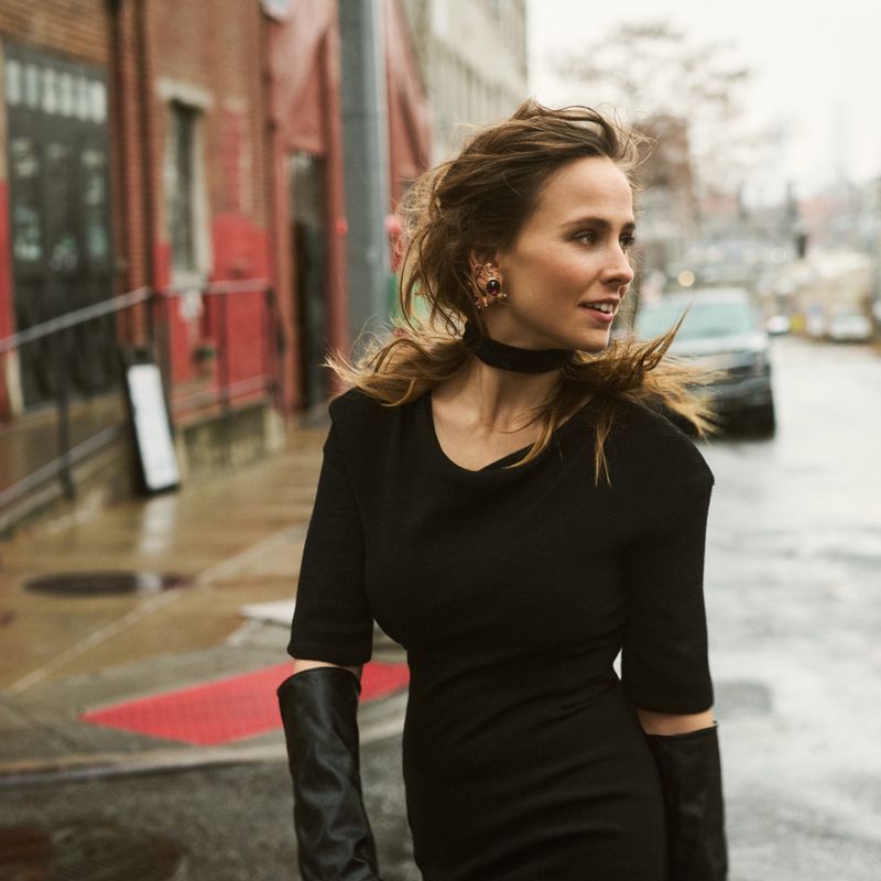 Adult woman in black dress poses on urban street. The woman is smiling and accessorized with earrings and gloves with neutral color grading.
