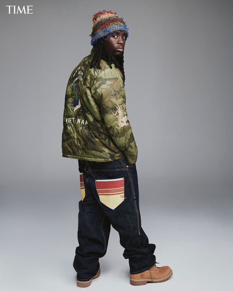 Full-length shot of a Black male model in a camouflage jacket, oversized jeans, tan boots, and a knitted beanie, standing against a gray background.