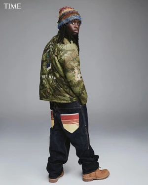 Kai Cenat wearing a camo jacket, knit cap, and wide-leg denim against a grey background for Time Magazine