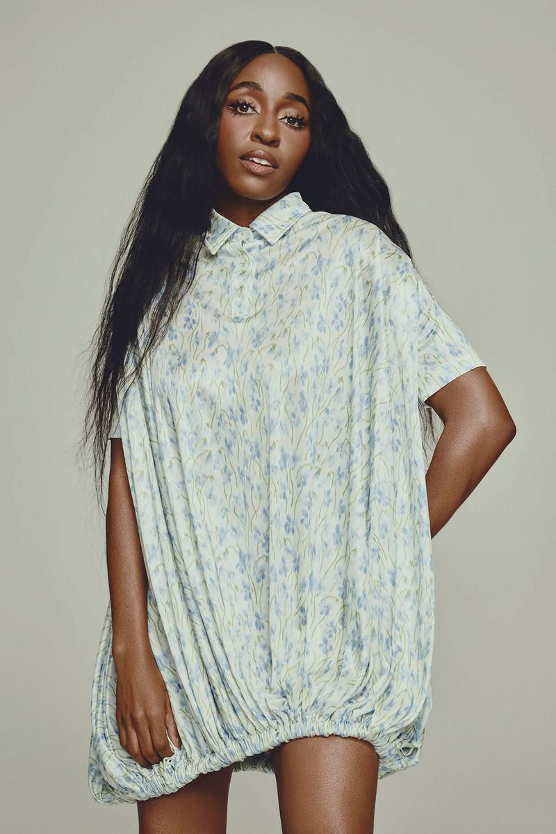 Portrait of a Black woman with long dark hair wearing a light blue patterned dress posing in a photo studio with a neutral backdrop.