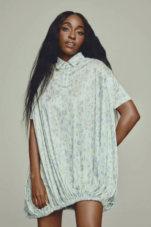 Portrait of a Black woman with long dark hair wearing a light blue patterned dress posing in a photo studio with a neutral backdrop.