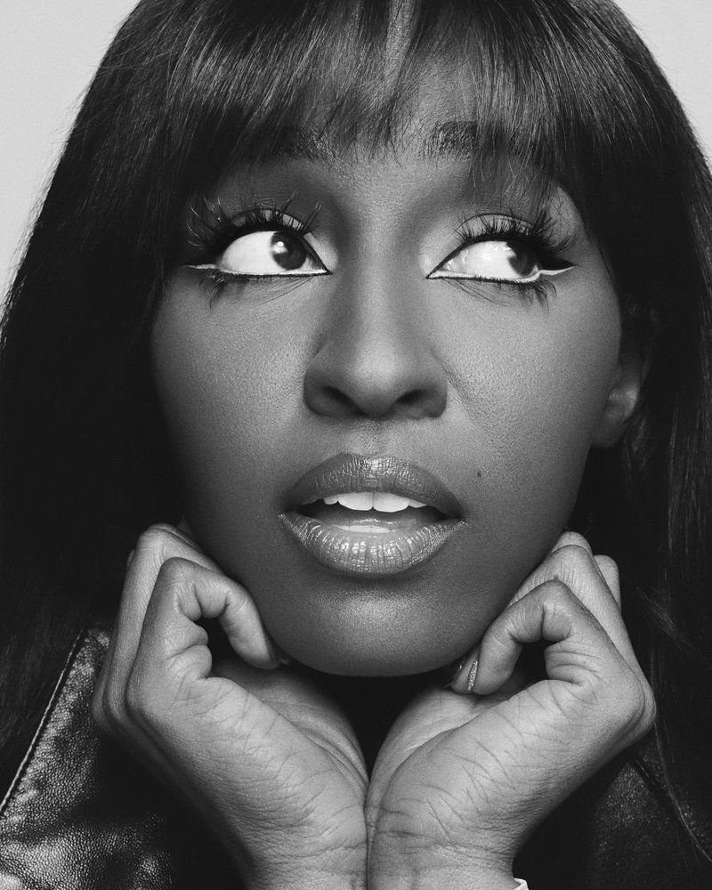 Black and white portrait of a Black woman with long dark hair and dramatic eye makeup looking up with hands near her face.