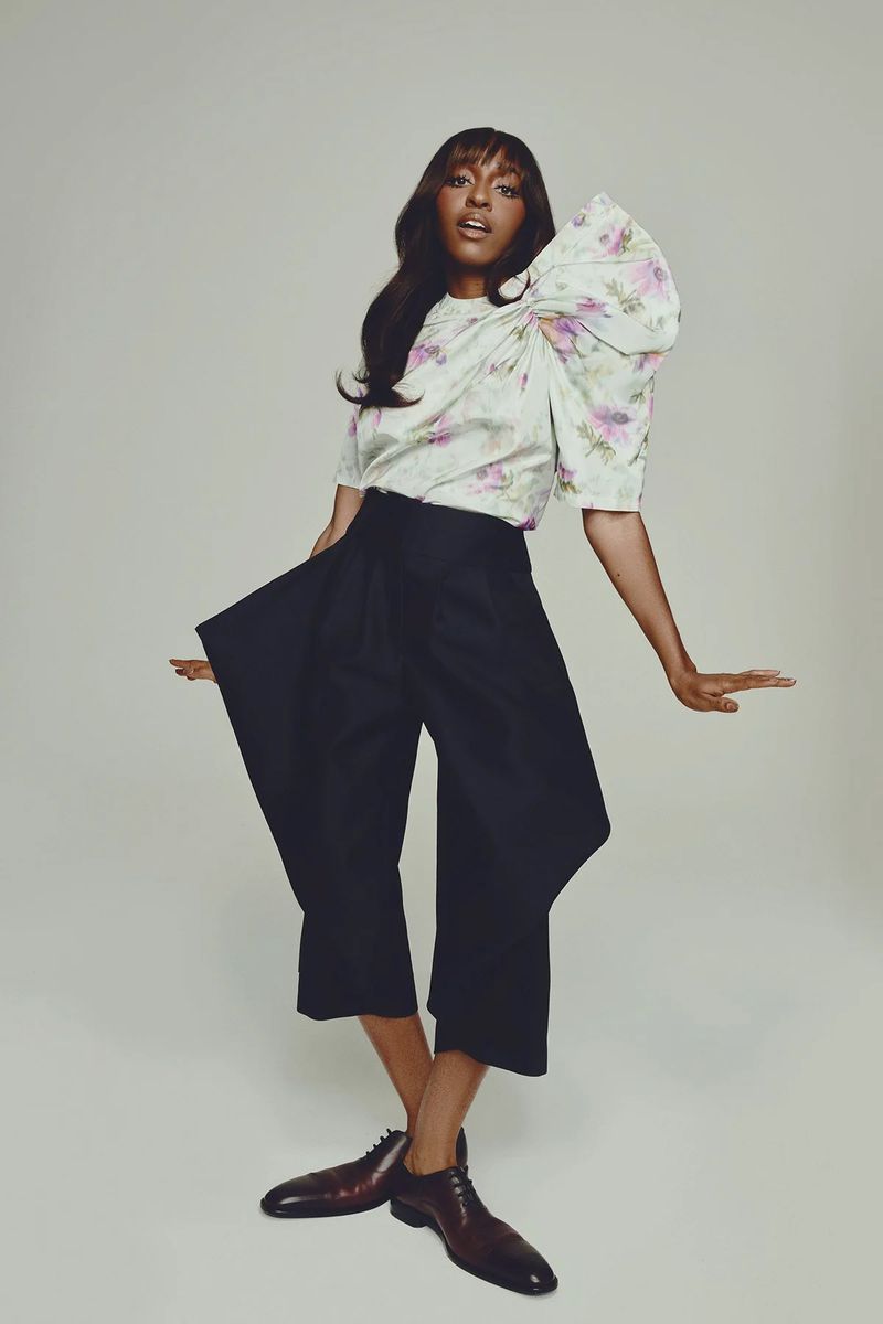 Fashionable woman in floral blouse and wide-leg trousers poses confidently in a studio with her arms out and legs crossed.