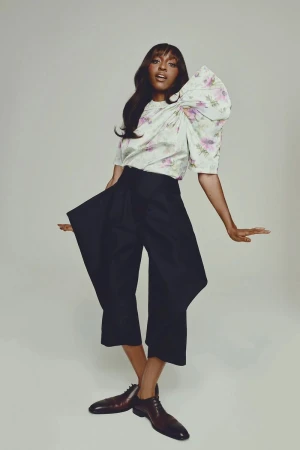 Fashionable woman in floral blouse and wide-leg trousers poses confidently in a studio with her arms out and legs crossed.