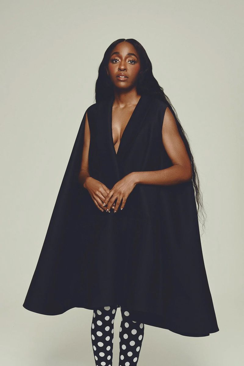 A striking portrait of a Black woman with long dark hair, wearing a sleeveless black dress and black and white polka dot tights.