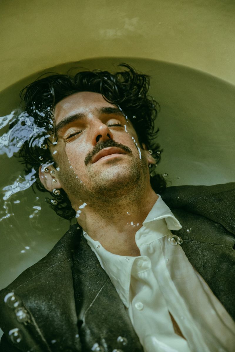 Adult male with a moustache is submerged in water, eyes closed, wearing a white shirt and dark blazer in an indoor setting.