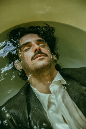 Adult male with a moustache is submerged in water, eyes closed, wearing a white shirt and dark blazer in an indoor setting.