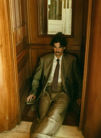 Young man with a mustache poses in a vintage-style suit in a wood-paneled interior with a neutral color palette and natural light.