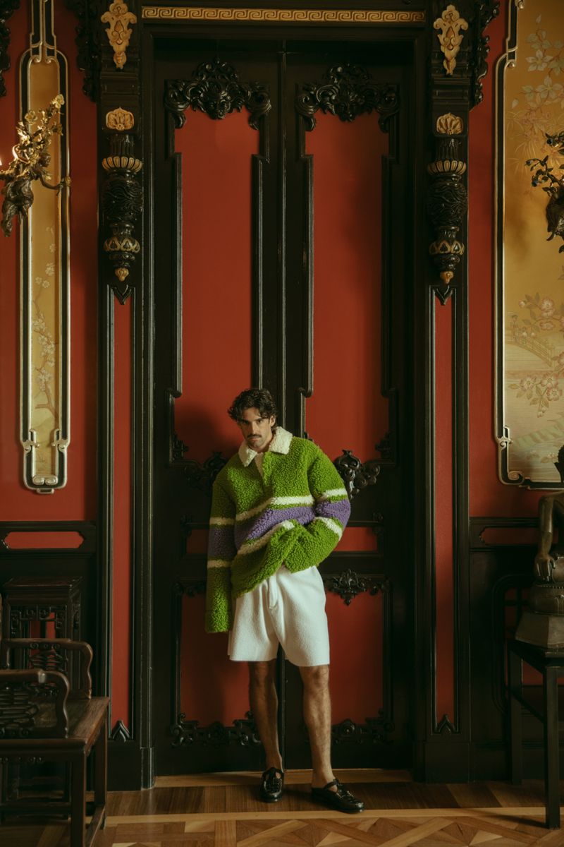 A stylish adult male stands in front of red double doors wearing a green sweater and white shorts in an ornate indoor setting.