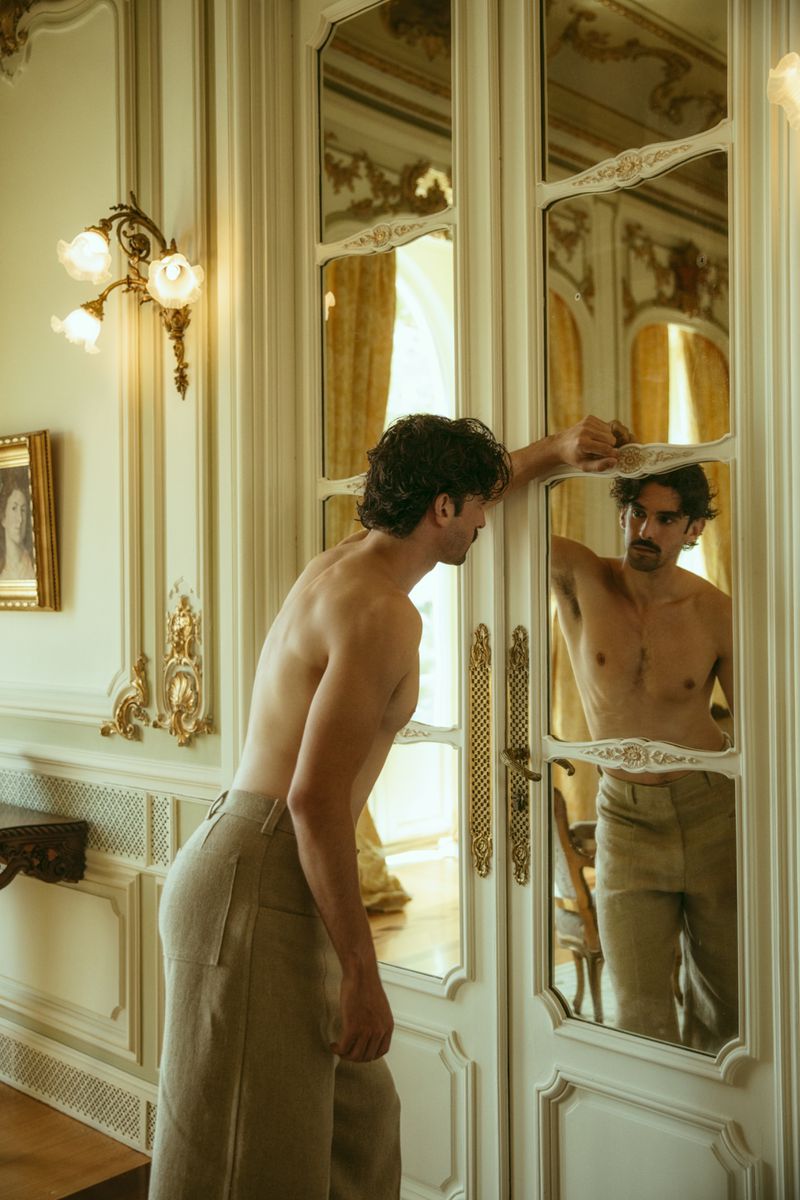 Shirtless man poses in front of a decorative wall mirror inside an ornately decorated room with neutral colors and natural lighting.