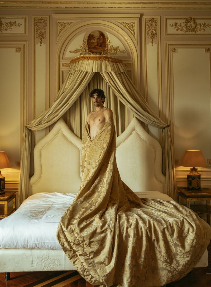 Adult male stands draped in gold jacquard textile in luxury hotel room with canopy bed, ornate wall panelling and parquet floors.