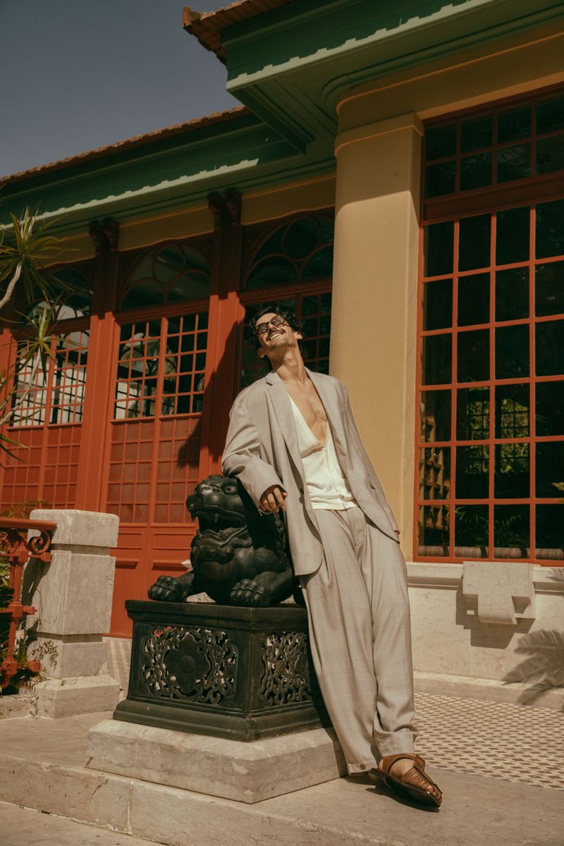 An adult male model in a light-grey suit leans against a decorative concrete lion statue near a building with red window frames outdoors.