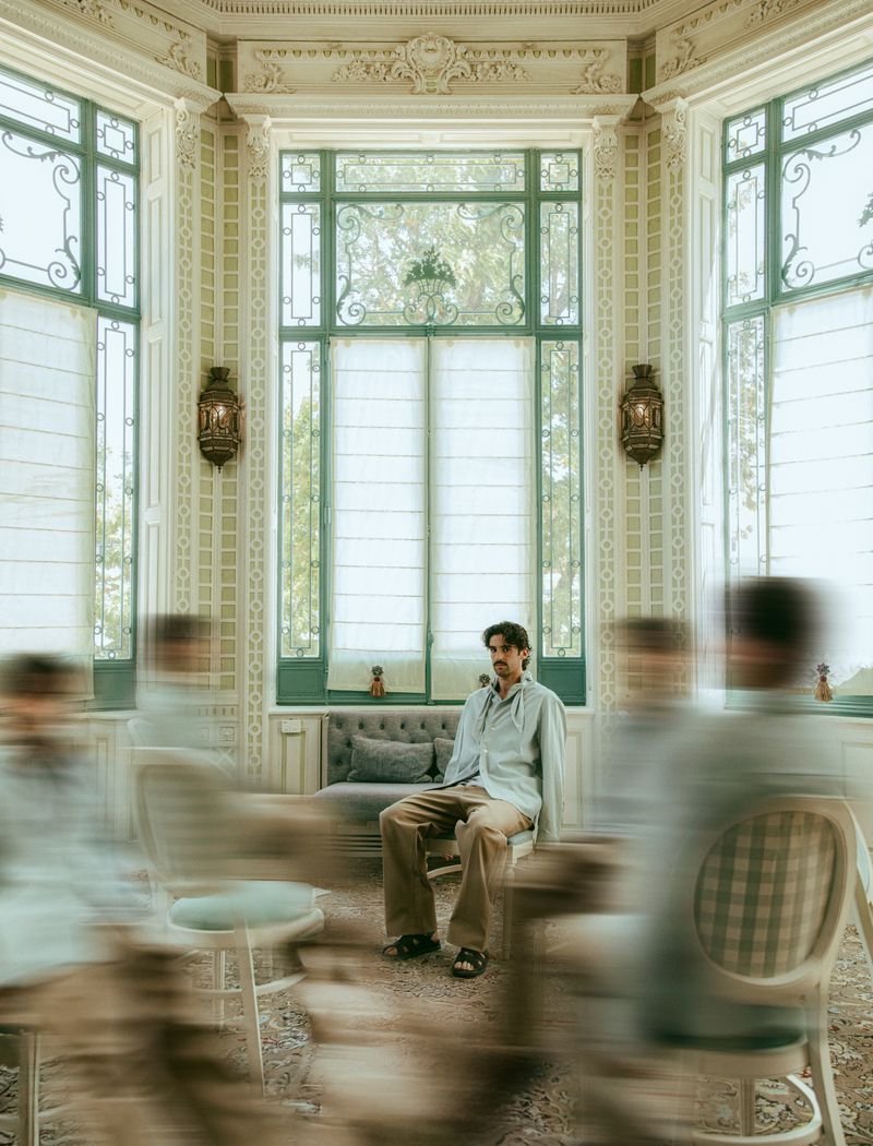 Adult male sitting in ornate room with large windows, wearing a blue shirt and tan pants. Blurred figures in motion surround him.