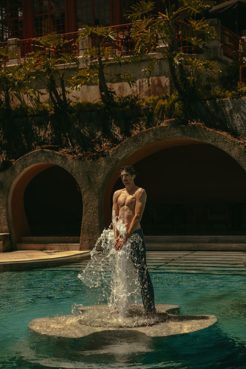 Shirtless man stands in swimming pool on stone platform with water flowing from his hands, with architectural backdrop and trees.